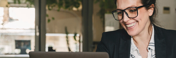 Financiacion a corto plazo / Liquidez - Mujer de negocios morena con gafas y camisa, sonriendo y trabajando frente a su portatil en la oficina